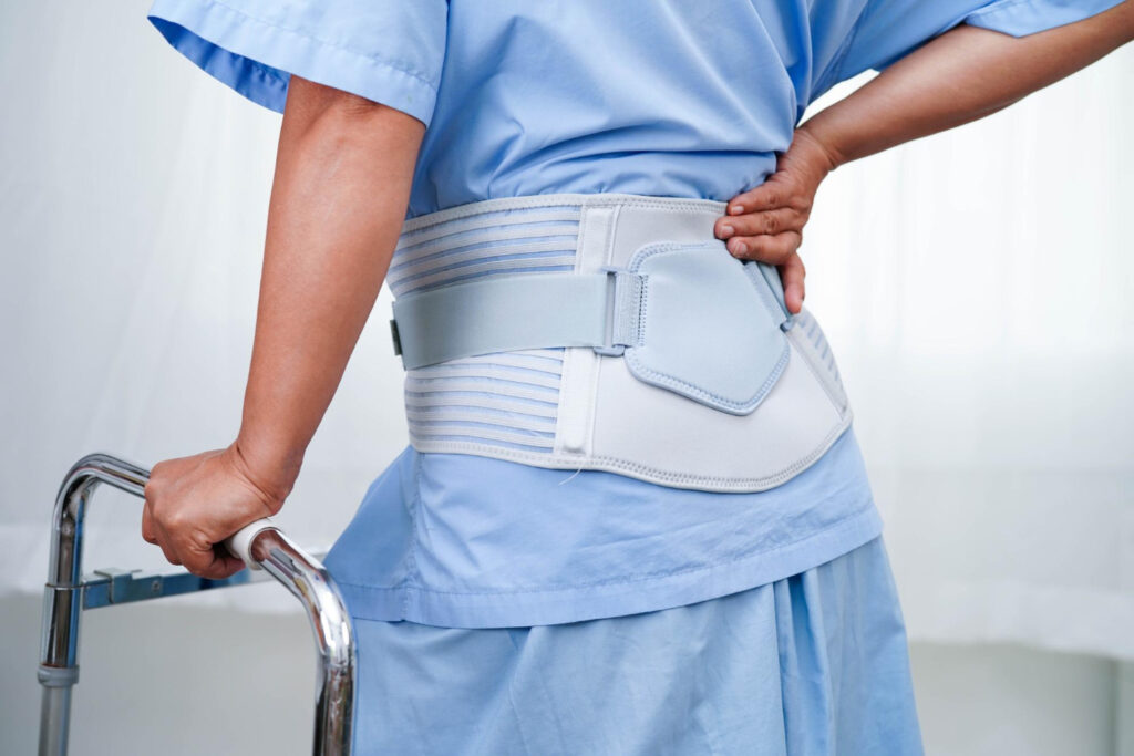 Person in a blue medical gown wearing a light gray lumbar back brace with straps, standing by a walker.