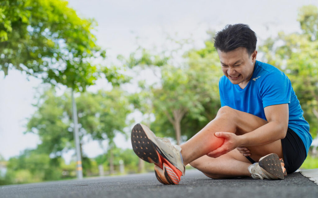 Man in a blue shirt sits on a paved path, grimacing in pain while holding his shin outdoors in a park setting