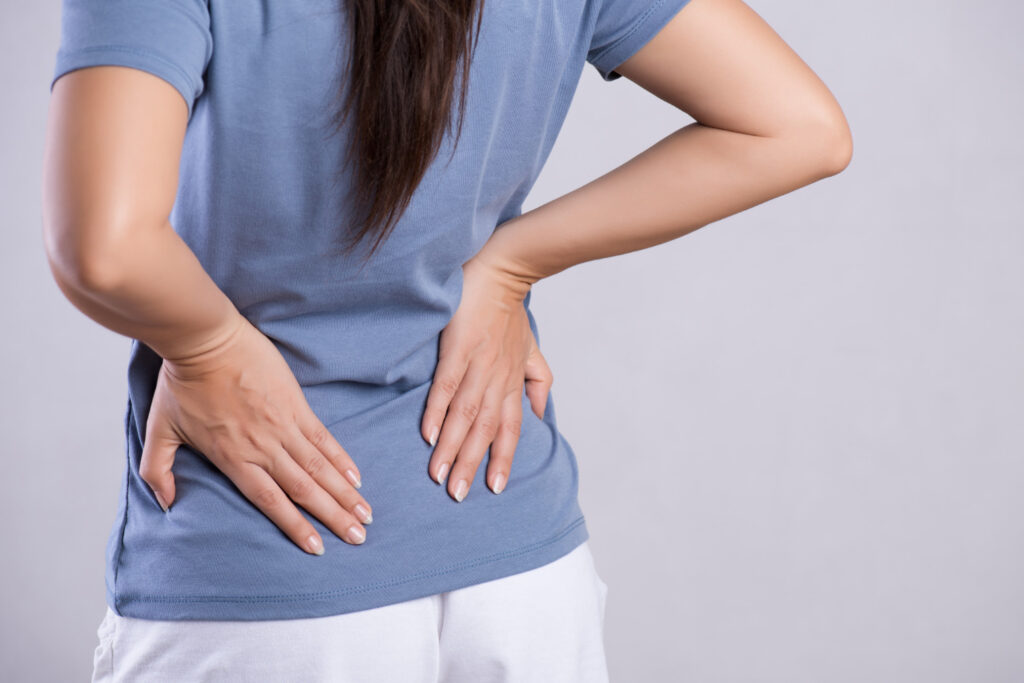 Person wearing a blue shirt holds their lower back with both hands, signaling back pain or discomfort.