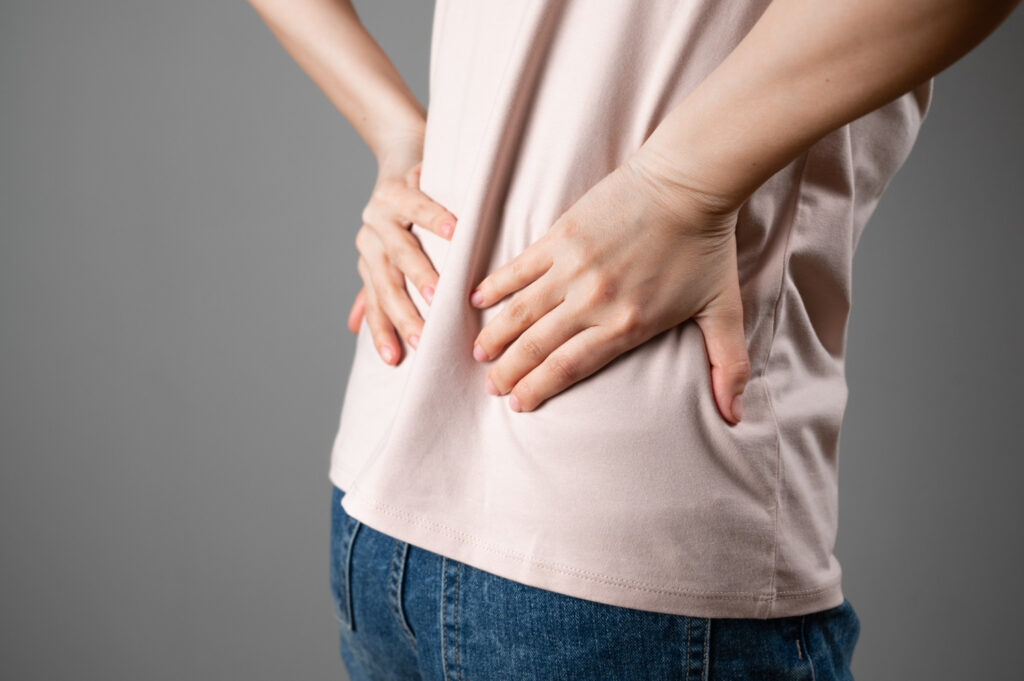 Person in a pale pink shirt stands with hands on the lower back, suggesting back pain or discomfort.