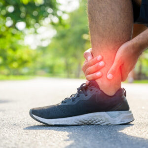 Person clutching a reddened ankle beside a black running shoe on a park path, indicating ankle pain or injury.