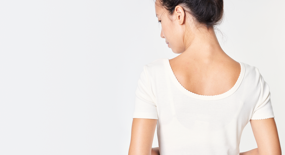 Back view of a woman with dark hair in a bun, wearing a white short-sleeve top, standing against a pale background