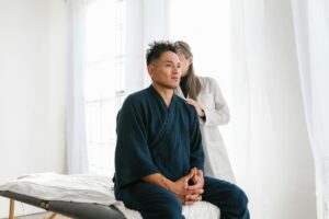 Man in a dark blue robe sits on an examination bed as a clinician in white coats his shoulders in a clinical setting.