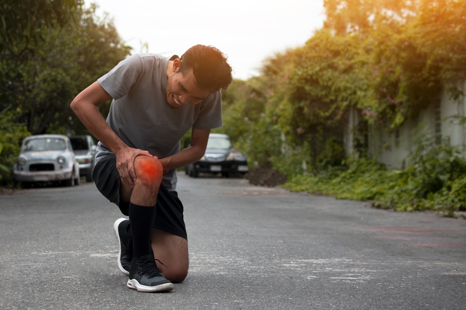 Man kneeling on a street, holding his injured knee with a grimace as sunlight glows from the right.